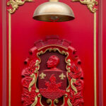 Blason rouge et or de la famille Chaudesaigues, situé sur la façade du restaurant Gourmet & Glouton à Chaudes-Aigues, représentant un visage sculpté et des ornements héraldiques.