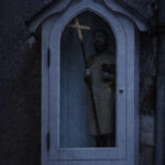 Niche religieuse ancienne à Chaudes-Aigues, avec statue tenant une croix, visible dans la pénombre sur une façade en pierre.