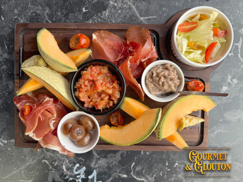 Vue du dessus de la planche estivale de Gourmet & Glouton à Chaudes-Aigues avec melon, jambon cru, tartare de tomates, caviar d'aubergines, artichauts, oignons Borettane et salade fraîche.