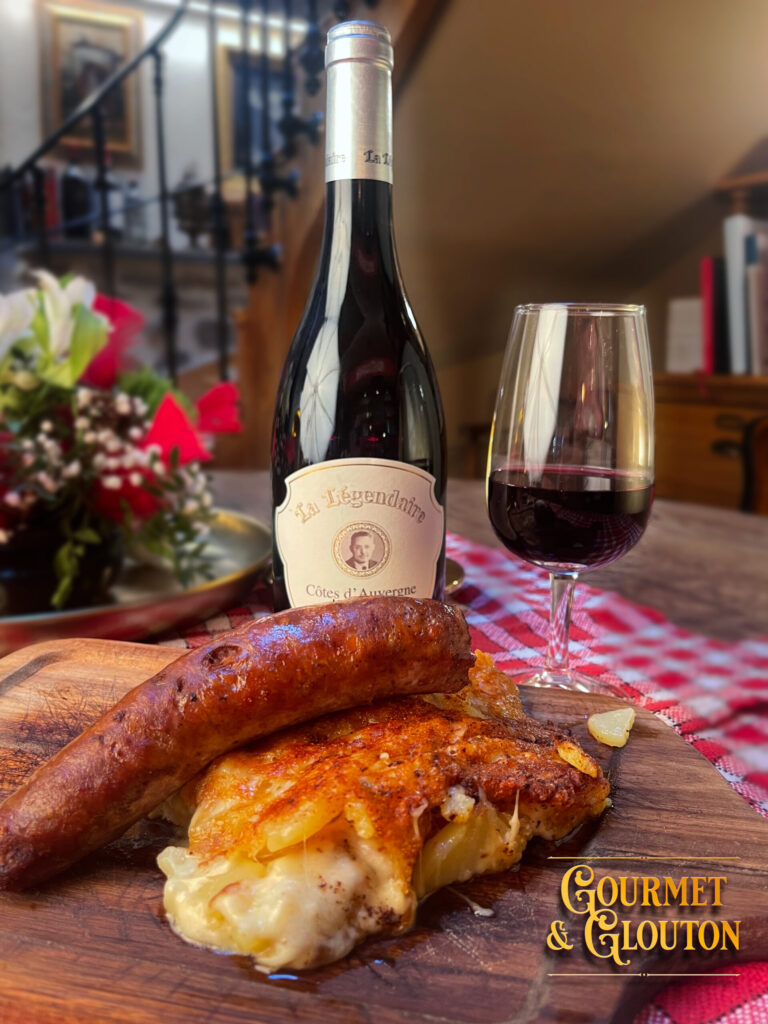 Saucisse maison sur une truffade dorée, avec une bouteille de La Légendaire Côtes d’Auvergne et un verre de vin rouge chez Gourmet & Glouton.
